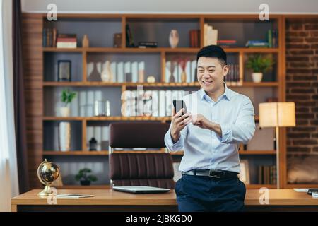 Homme d'affaires asiatique prospère dans un bureau à domicile moderne, regarde l'écran du téléphone, sourit et se réjouit, portrait de indépendant réussi en chemise, utilise le téléphone Banque D'Images