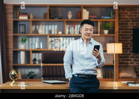 Homme d'affaires asiatique prospère dans un bureau à domicile moderne, regarde l'écran du téléphone, sourit et se réjouit, portrait de indépendant réussi en chemise, utilise le téléphone Banque D'Images