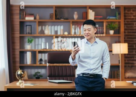 Homme d'affaires asiatique prospère dans un bureau à domicile moderne, regarde l'écran du téléphone, sourit et se réjouit, portrait de indépendant réussi en chemise, utilise le téléphone Banque D'Images