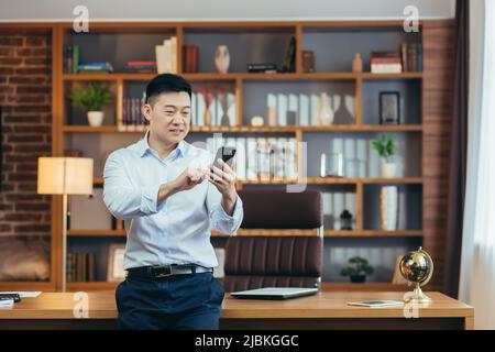 Homme d'affaires asiatique prospère dans un bureau à domicile moderne, regarde l'écran du téléphone, sourit et se réjouit, portrait de indépendant réussi en chemise, utilise le téléphone Banque D'Images