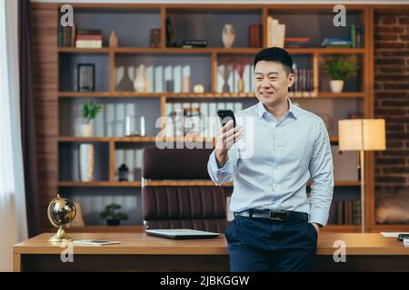 Homme d'affaires asiatique prospère dans un bureau à domicile moderne, regarde l'écran du téléphone, sourit et se réjouit, portrait de indépendant réussi en chemise, utilise le téléphone Banque D'Images