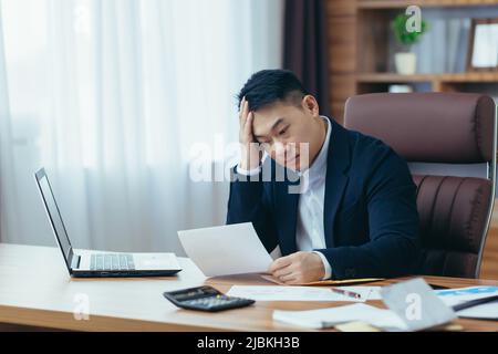 Jeune homme asiatique comptable, employé de bureau travaillant avec des documents, fatigué, assis à un bureau dans le bureau Banque D'Images