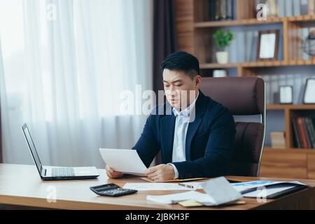 Comptable-auditeur travaille dans un bureau classique, calcule les rapports financiers, homme d'affaires asiatique travaille avec l'ordinateur et les documents, la paperasse Banque D'Images