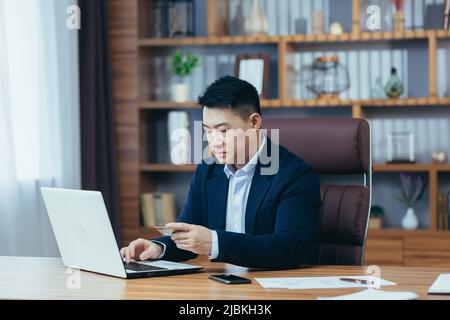 Un homme d'affaires asiatique prospère effectue des opérations bancaires et transfère de l'argent à partir d'un compte électronique, utilise un ordinateur portable et une carte de crédit bancaire Banque D'Images