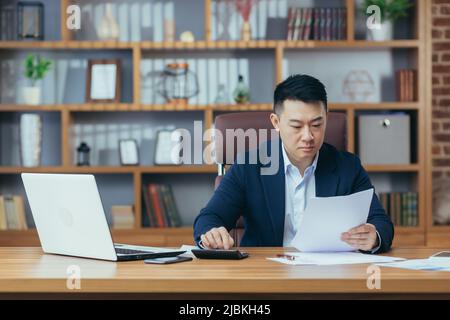 Comptable-auditeur travaille dans un bureau classique, calcule les rapports financiers, homme d'affaires asiatique travaille avec l'ordinateur et les documents, la paperasse Banque D'Images