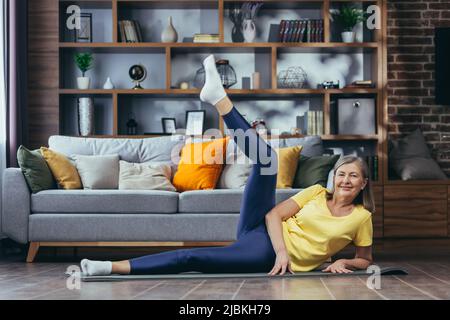 Femme sénior aux cheveux gris faisant de la forme physique à la maison, faisant des exercices matinaux sur un tapis de sport assis sur le sol à la maison Banque D'Images