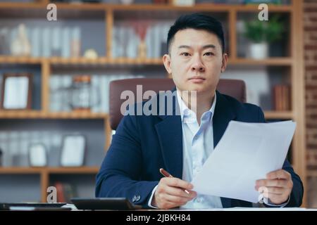 Portrait d'un jeune homme d'affaires asiatique beau, réalisateur travaillant avec des documents au bureau, sérieux, regardant l'appareil photo Banque D'Images