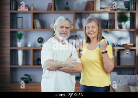 Couple marié retraité senior faisant de la forme physique à la maison, debout dans la salle de séjour avec des haltères et souriant regardant l'appareil photo Banque D'Images