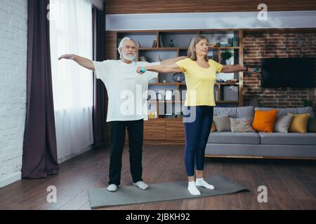 Couple marié retraité senior faisant de la forme physique à la maison, debout dans la salle de séjour avec des haltères et souriant Banque D'Images