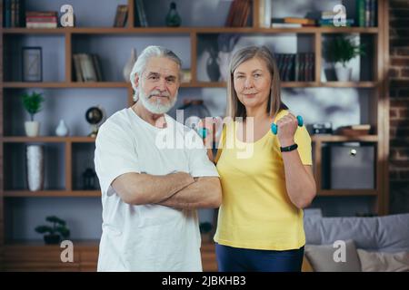 Couple marié retraité senior faisant de la forme physique à la maison, debout dans la salle de séjour avec des haltères et souriant regardant l'appareil photo Banque D'Images