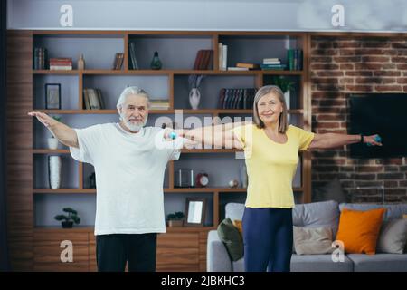 Couple marié retraité senior faisant de la forme physique à la maison, debout dans la salle de séjour avec des haltères et souriant regardant l'appareil photo Banque D'Images