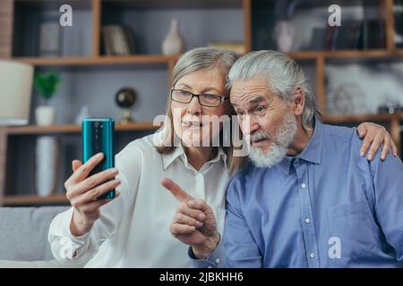 Couple de retraités âgés heureux grands-parents gris cheveux communiquent en ligne dans un appel vidéo à l'aide d'un téléphone portable smartphone. Rire et vague ha Banque D'Images