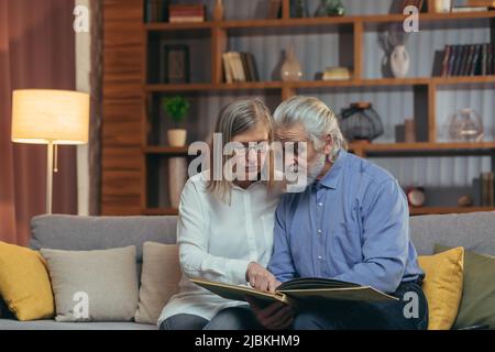 Un charmant couple senior regardant l'album photo Memoirs sur le canapé à la maison. Grands-parents de famille âgés assis ensemble regardant et discutant agréable mome Banque D'Images