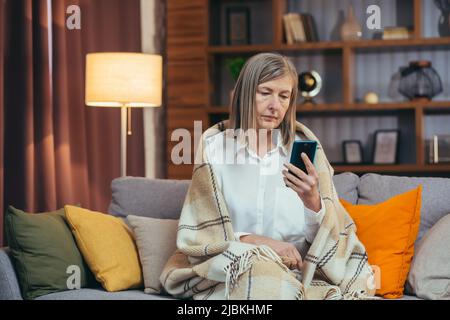 Femme sénior aux cheveux gris à la maison assise sur un canapé et regardant l'écran du téléphone Banque D'Images