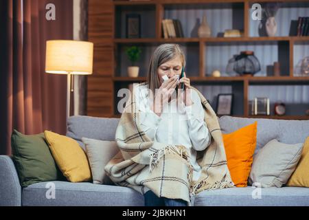 Une femme malade aux cheveux gris à la maison, a un nez qui coule, s'assoit sur le canapé, appelle le médecin pour obtenir de l'aide et des conseils Banque D'Images