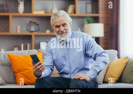 Un vieil homme heureux utilise le téléphone à la maison, assis sur le canapé, utilise des applications mobiles Banque D'Images