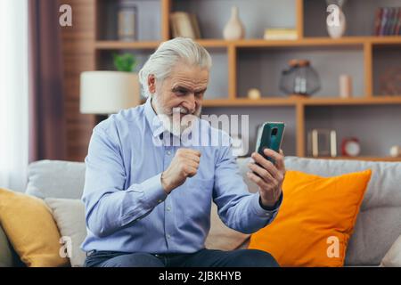 Un vieil homme heureux utilise le téléphone à la maison, assis sur le canapé, utilise des applications mobiles Banque D'Images