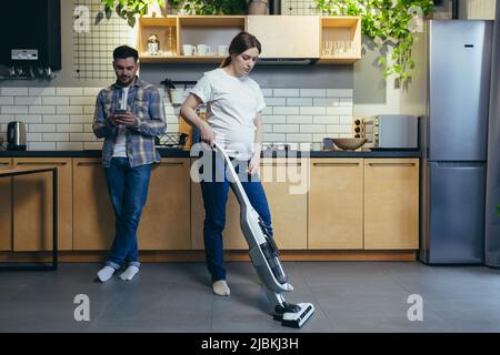 Famille à la maison, femme enceinte nettoie la pièce avec un aspirateur, mari utilise le téléphone dans la cuisine Banque D'Images