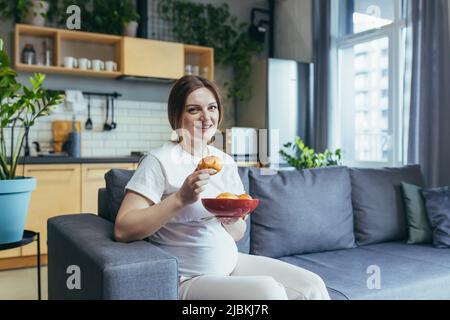 Femme enceinte à la maison assise sur un canapé mangeant des beignets malsains et de la malbouffe Banque D'Images