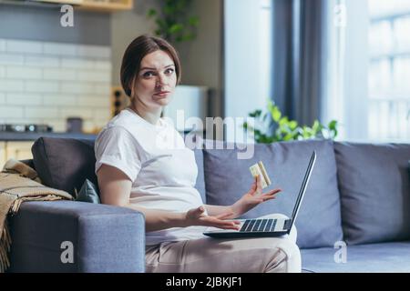 Une femme enceinte frustrée essaie de faire des achats en ligne à la maison, regarde la caméra dans la frustration, utilise un ordinateur portable, tient une carte de crédit bancaire Banque D'Images