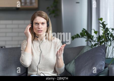 Une femme en colère qui parle lors d'un appel vidéo regarde la caméra et explique qu'elle utilise un casque, travaille à distance depuis sa maison, assis sur son canapé Banque D'Images