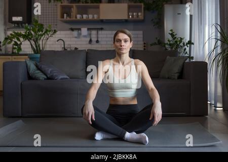 Heureuse femme d'âge moyen assise dans la position de lotus méditant, faisant de la forme physique le matin à la maison, regardant l'appareil photo Banque D'Images