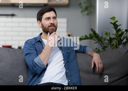 L'homme à la maison est assis sur le canapé, se reposant en regardant loin, souriant pensif de rêve Banque D'Images