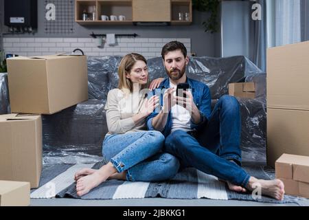 Un homme et une femme dans un nouvel appartement, un couple marié assis sur le sol, près des boîtes en carton, en utilisant le téléphone pour les achats en ligne, en choisissant des meubles Banque D'Images