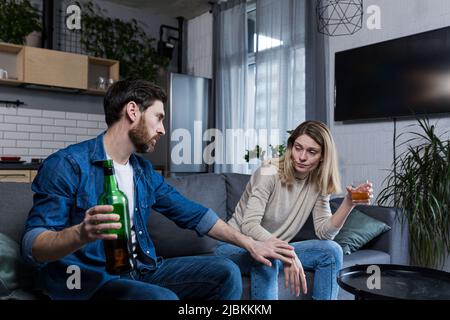 Alcoolisme féminin, querelle familiale à la maison, femme buvant de l'alcool, mari essayant de l'arrêter, assis sur le canapé Banque D'Images