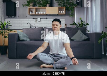 Asiatique lui-même médite à la maison le matin, assis sur un tapis de sport, près du canapé dans le salon, son matin, calme à l'extérieur du lotus Banque D'Images