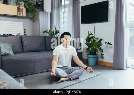 Asiatique lui-même médite à la maison le matin, assis sur un tapis de sport, près du canapé dans le salon, son matin, calme à l'extérieur du lotus Banque D'Images