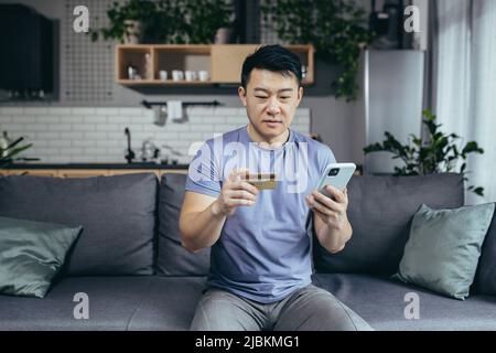 homme seul à la maison, shopping en ligne, asiatique utilise le téléphone et l'application d'achat, détient une carte de crédit, est assis sur un canapé à la maison Banque D'Images