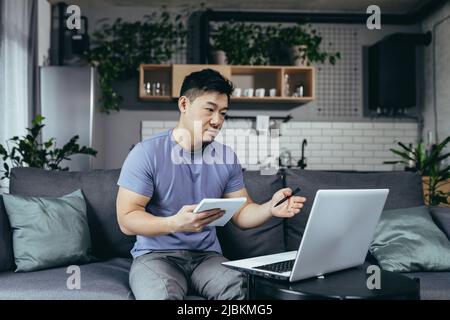 Un homme étudie à la maison, utilise un ordinateur portable pour l'apprentissage en ligne, un asiatique s'assoit sur le canapé, écrit de nouvelles informations Banque D'Images