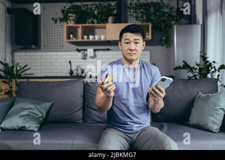 homme seul à la maison, shopping en ligne, asiatique utilise le téléphone et l'application d'achat, détient une carte de crédit, est assis sur un canapé à la maison Banque D'Images