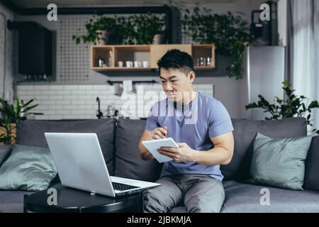 Un homme étudie à la maison, utilise un ordinateur portable pour l'apprentissage en ligne, un asiatique s'assoit sur le canapé, écrit de nouvelles informations Banque D'Images