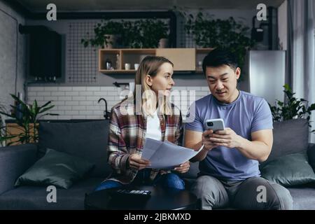 papier de devoirs, couple multiracial, homme et femme, payer des factures par téléphone et application, asseyez-vous à la maison sur le canapé, heureux et souriant Banque D'Images