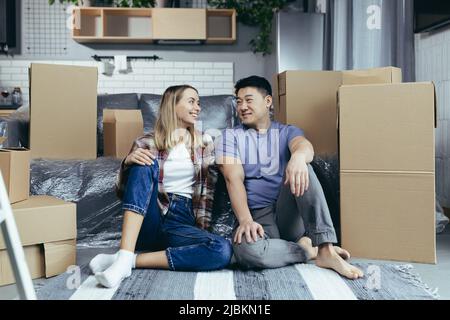 Jeune couple multiracial asiatique homme et femme, dans un appartement nouvellement loué, s'embrassant assis sur le plancher de la maison, boîtes en carton déballé Banque D'Images