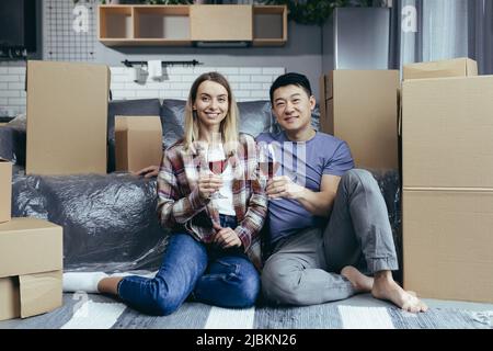 Jeune heureux couple de boire du vin dans des verres assis sur le sol dans un nouvel appartement, asiatique homme et femme heureux ensemble, parmi des boîtes en carton, juste m Banque D'Images