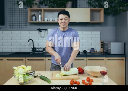 L'homme est seul à la maison, préparant une salade, trancher des légumes dans la cuisine, l'asiatique regarde l'appareil photo et sourit Banque D'Images