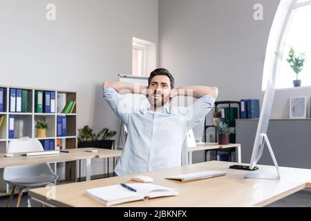 Jeune homme beau, employé de bureau, assis à un bureau d'ordinateur, étirant, fatigué, prenant une pause, faisant des exercices de yoga, repos Banque D'Images