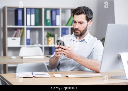 Jeune homme concentré avec une barbe, utilise le téléphone, assis à la maison à la table, lit soigneusement de l'écran Banque D'Images