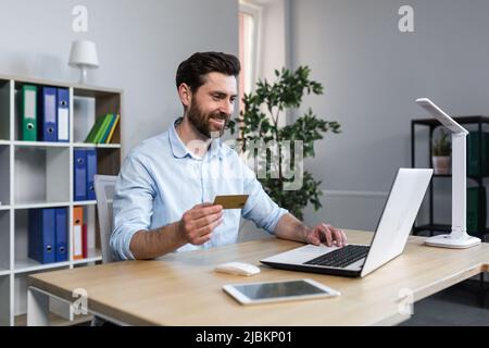 Un homme d'affaires heureux fait des achats en ligne travaillant dans le bureau, le mari tient la carte de crédit et utilise l'ordinateur portable Banque D'Images