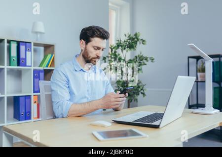 Jeune homme concentré avec une barbe, utilise le téléphone, assis à la maison à la table, lit soigneusement de l'écran Banque D'Images