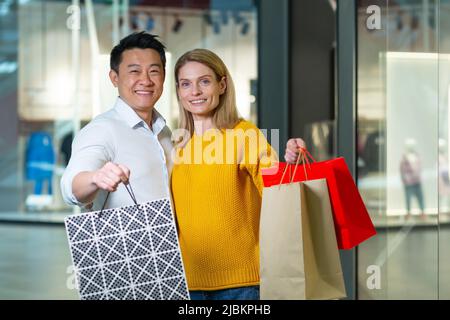 Un jeune homme et une jeune femme asiatiques se coupent dans les magasins de supermarché, regardant la caméra et souriant, tenant des paquets colorés avec des marchandises Banque D'Images