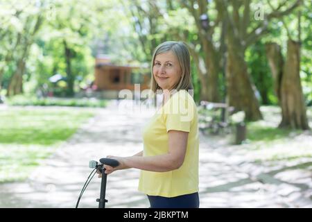 Portrait d'une femme active senior, d'une femme aux cheveux gris dans le parc, regardant l'appareil photo et souriant à vélo, jour d'été Banque D'Images