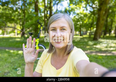 La femme sénior, active sportswoman dans le parc en été, regarde la caméra du téléphone et sourit en lui agitant la main lors d'un appel vidéo Banque D'Images