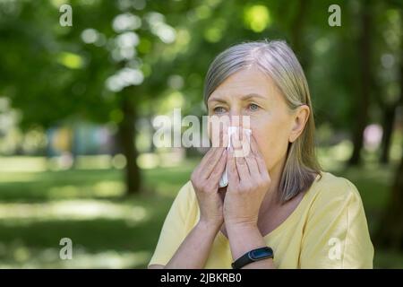 Une femme âgée dans le parc qui souffre d'allergies a un nez qui coule et éternue près des arbres Banque D'Images