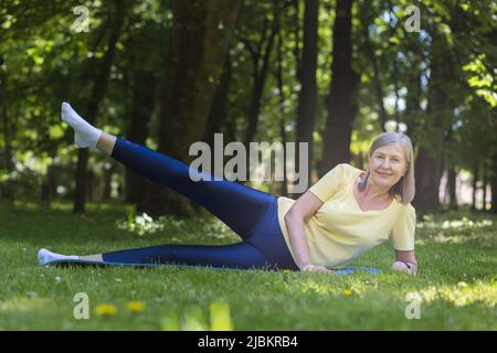 Un mode de vie actif et sain. Portrait d'une belle femme âgée. Effectue des exercices de sport, allongé dans le parc sur l'herbe sur un tapis de sport, regardant Banque D'Images