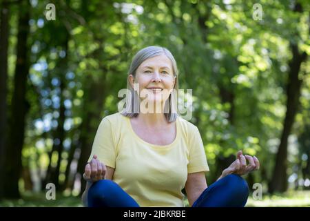 Un mode de vie actif et sain. Portrait d'une belle femme âgée. Il pratique le yoga. Elle est assise dans le parc sur l'herbe sur un tapis de sport, regardant le Banque D'Images
