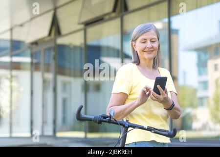 La femme âgée aux cheveux gris aime le téléphone lors d'une promenade estivale avec un vélo, pensionné souriant Banque D'Images